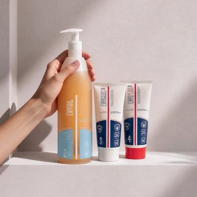 Mano che tiene l'olio detergente Envioil® accanto a due creme grasse per pelle secca atopica